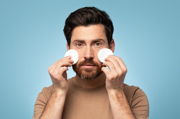 Bel homme d'âge moyen nettoyant la peau du visage avec des tampons de coton et regardant la caméra sur fond bleu