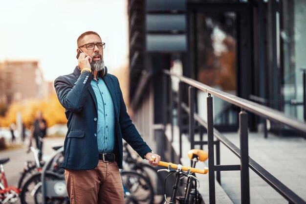 Un bel homme d'âge moyen se rend au travail à vélo, attend quelqu'un et parle sur le smartphone devant le quartier des bureaux.