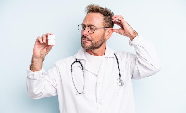 Bel Homme D'âge Moyen Se Sentant Perplexe Et Confus, Se Grattant La Tête. Médecin Avec Bouteille De Pilules