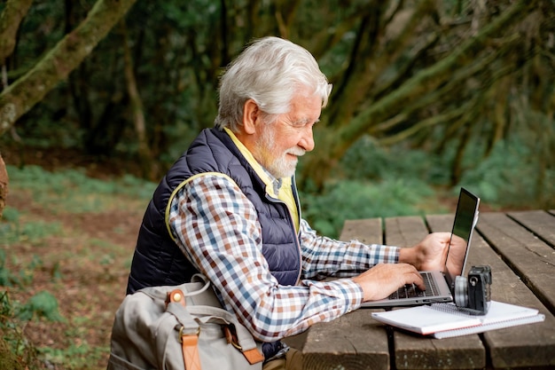 Bel homme âgé souriant travaillant sur un ordinateur portable tout en étant assis à la table en bois à l'extérieur dans le parc