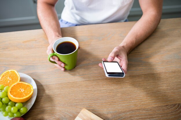 Bel homme à l&#39;aide de smartphone et tenant la tasse dans la cuisine