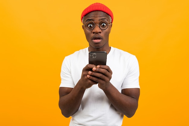 Bel homme américain dans un T-shirt blanc avec un chapeau rouge communique sur le réseau social dans un smartphone avec un visage surpris