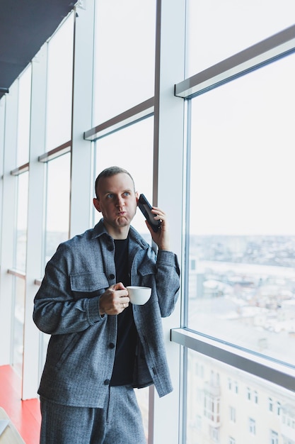 Un bel homme d'apparence européenne dans un costume décontracté boit du café parle au téléphone et regarde par la fenêtre