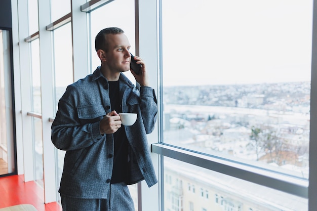 Un bel homme d'apparence européenne dans un costume décontracté boit du café parle au téléphone et regarde par la fenêtre