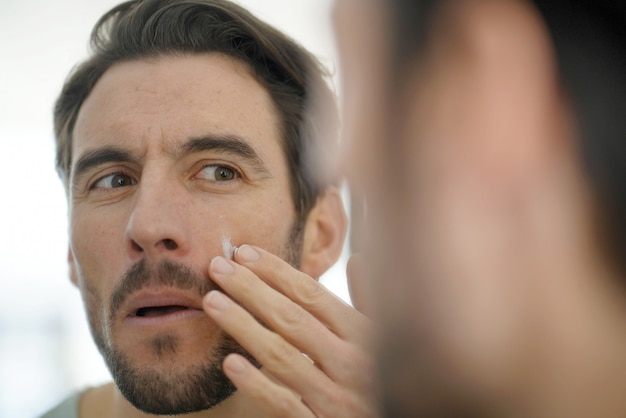 Bel homme, appliquer, crème visage, regarder dans miroir