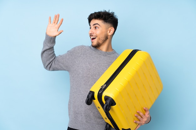 Bel homme arabe isolé sur mur bleu en vacances avec valise de voyage et saluant