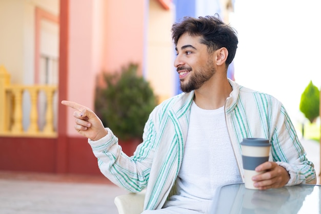 Bel homme arabe tenant un café à emporter à l'extérieur pointant vers le côté pour présenter un produit