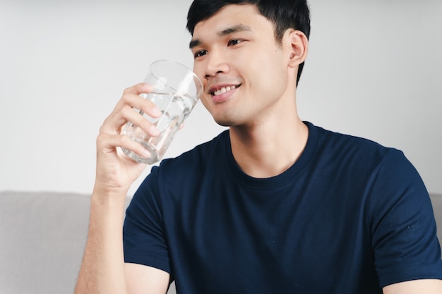 Bel homme asiatique buvant un verre d'eau sur le canapé du salon