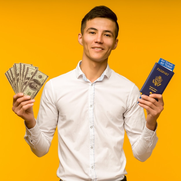 Un bel homme asiatique en chemise blanche se réjouit d'avoir remporté la loterie. Il tient un passeport avec des billets d'avion et des dollars sur un espace jaune.