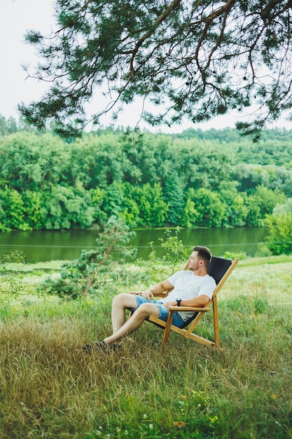 Bel homme assis sur une chaise en bois seul dans la nature Jeune bel homme assis sur un banc à l'ombre des arbres et profitant de la nature environnante par une journée ensoleillée