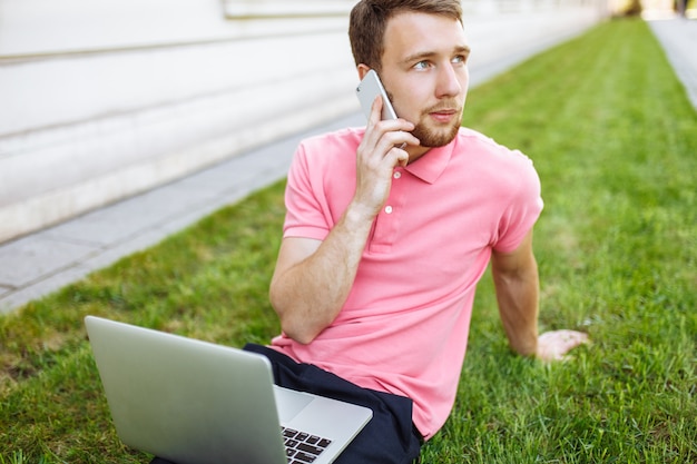 Bel homme assis sur l'herbe dans la ville avec un ordinateur portable et parler au téléphone, recherche d'emploi