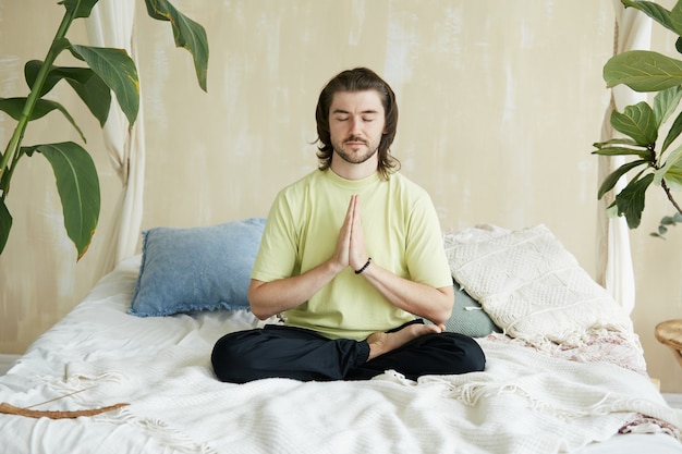Bel homme assis sur le lit en posture de lotus et namaste, conscience et concept d'yoga, espace copie