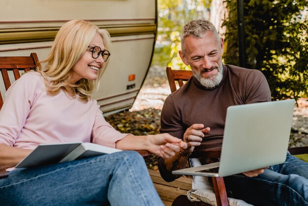 Un bel homme aux cheveux gris mature montre à sa femme blonde quelque chose d'amusant sur son ordinateur portable dans leur jardin.