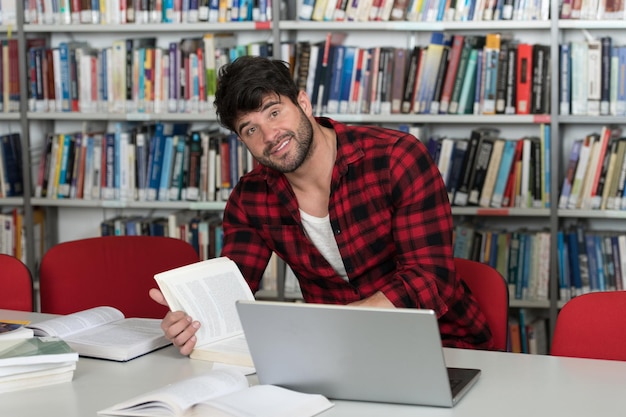 Bel homme aux cheveux noirs assis à un bureau dans la bibliothèque Ordinateur portable et organisateur sur la table en regardant l'écran un concept d'étude de livres flous à l'arrière