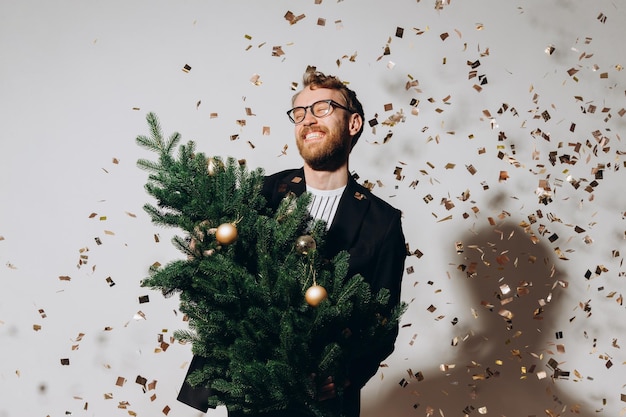 Bel homme aux cheveux roux dans une veste noire et des lunettes danse avec un arbre de Noël dans ses mains Concept de fête du Nouvel An