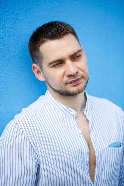 Un bel homme avec une barbe dans une chemise se dresse sur un fond bleu.