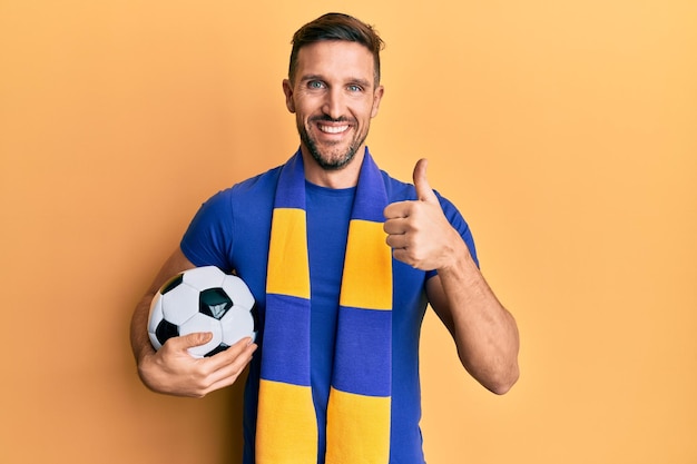 Bel homme avec barbe football hooligan jeu d'encouragement tenant le ballon souriant heureux et positif, pouce vers le haut faisant un excellent signe d'approbation