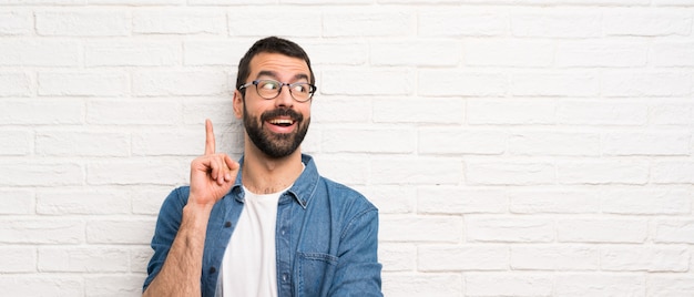 Bel homme à la barbe sur le mur de briques blanches pensant une idée pointant le doigt vers le haut