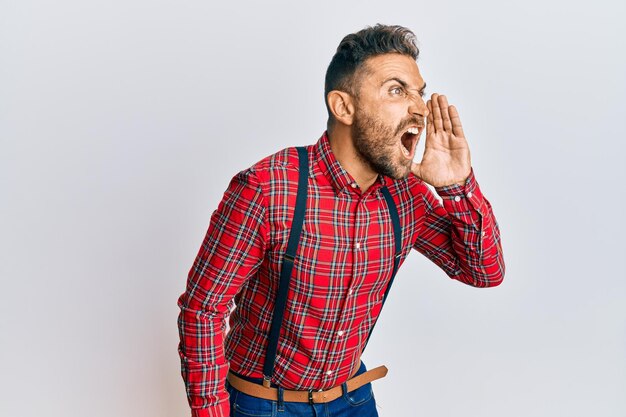 Bel homme avec barbe portant un look élégant hipster criant et hurlant fort à côté avec la main sur la bouche. notion de communication.