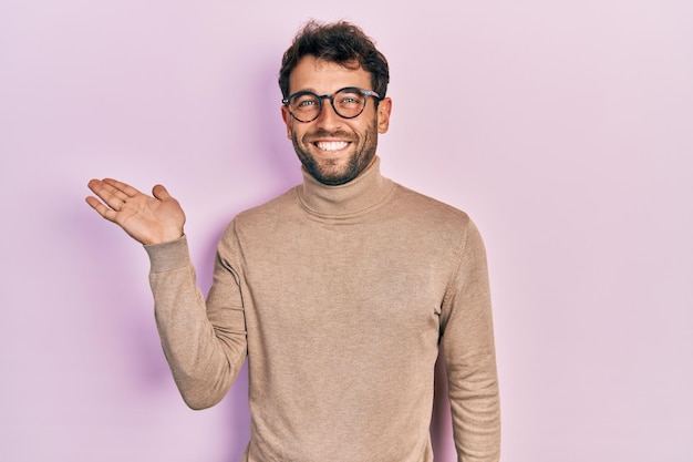 Bel homme avec barbe portant un pull à col roulé et des lunettes souriant joyeux présentant et pointant avec la paume de la main en regardant la caméra