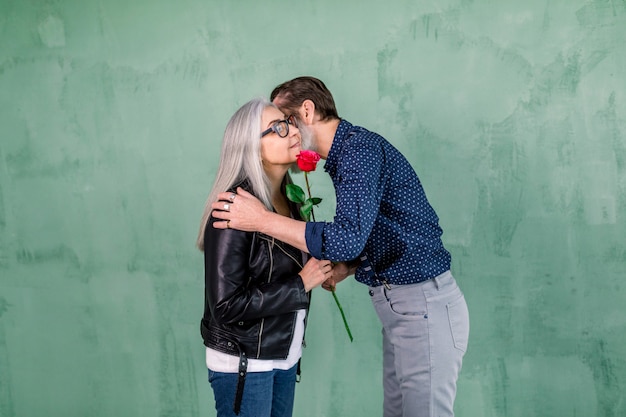 Bel homme barbu âgé donnant une belle rose fraîche à sa charmante femme souriante et embrassant sa joue, debout sur le mur vert