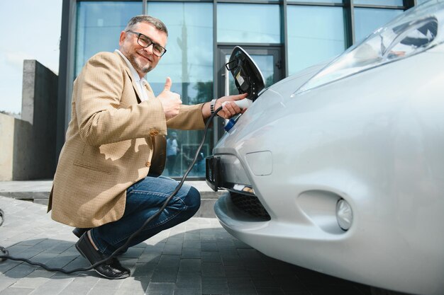 Bel homme barbu assis près de sa nouvelle voiture électrique moderne et tenant la prise du chargeur pendant que la voiture se charge à la station de charge