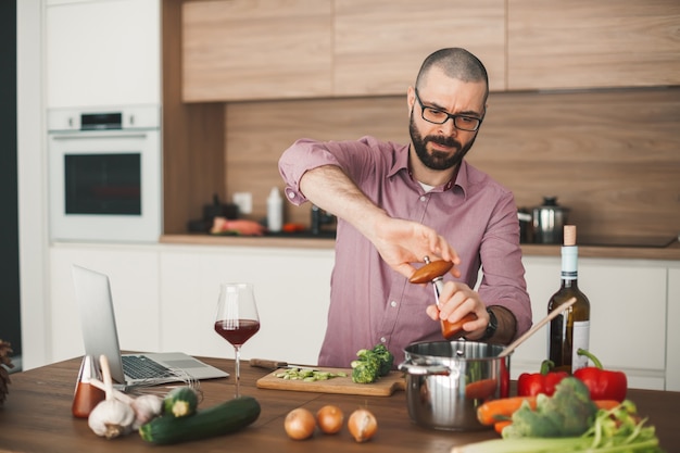 Bel homme barbu, cuisson du ragoût de légumes à l'aide d'un ordinateur portable. Il coupe un brocoli, une courgette, un poivron rouge, un oignon et d'autres légumes.