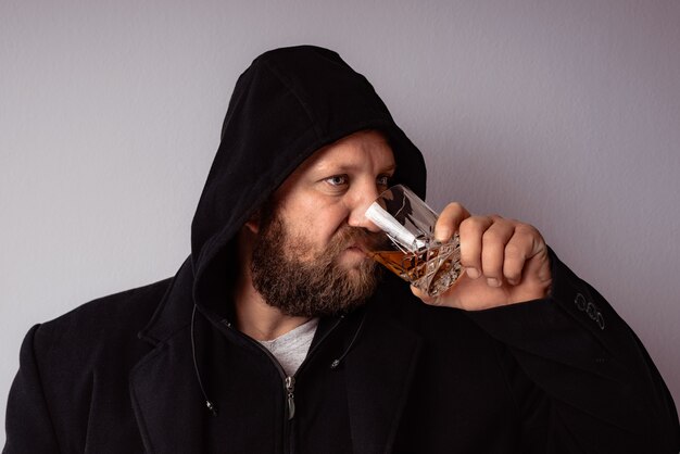 Bel homme barbu élégant portant un manteau noir avec capuche boisson avec de l'alcool fort