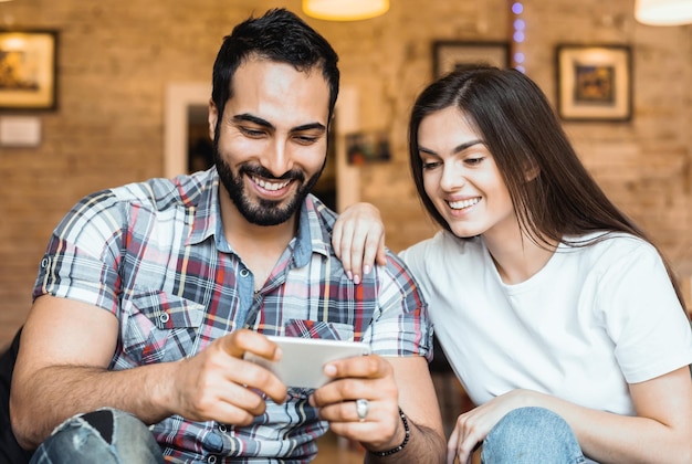 Bel homme barbu et fille brune s'amusant à jouer à un jeu en ligne sur smartphone dans une chambre élégante