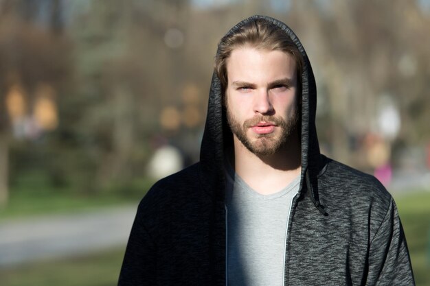 Bel homme barbu ou grave gars à capuchon avec barbe ensoleillée en plein air sur fond flou