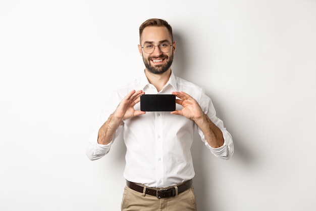 Bel homme barbu à lunettes, démontrant une application de téléphonie mobile, montrant l'écran du smartphone, debout sur fond blanc.