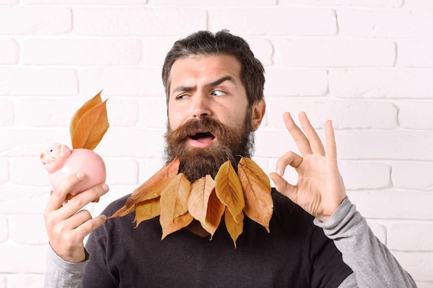 Bel homme barbu avec une moustache élégante et des feuilles jaunes d'automne dans une longue barbe sur un visage drôle avec une tirelire rose montrant ok sur fond de mur de briques blanches