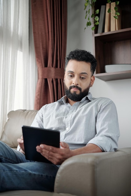 Bel homme barbu positif assis dans un fauteuil à la maison et lisant un livre ou un article sur tablette comp...