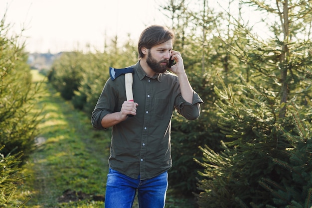 Bel homme barbu positif avec hache qui marche parmi les rangées d'arbres de Noël dans la foresterie et