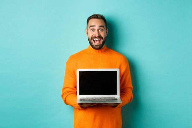 Bel homme barbu en pull orange montrant un écran d'ordinateur portable, démontrant une promo, debout sur fond bleu clair