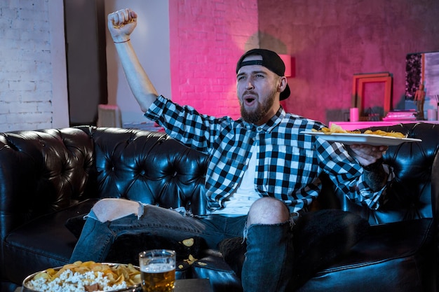 Un bel homme barbu regarde un match de football à la télévision assis à la maison sur le canapé et boit de la bière.