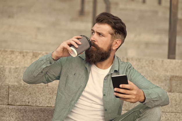 Bel homme barbu relaxant téléphone portable et tasse de café Vivre la vie en ligne Réseaux sociaux Communication en ligne de la vie moderne Mec avec fond d'espace urbain smartphone Technologie moderne