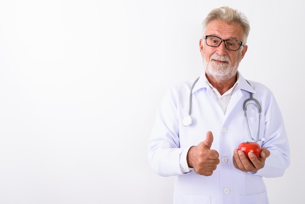 Bel homme barbu senior médecin tenant la tomate rouge tout en abandonnant le pouce sur blanc