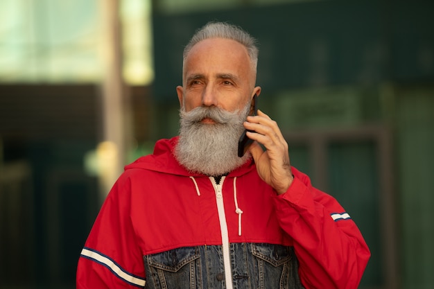 Bel homme barbu senior parle par un smartphone tout en prenant une pause-café dans une rue à côté de l'immeuble de bureaux.