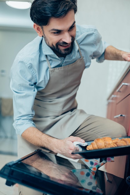 Bel homme barbu souriant et regardant les croissants en les sortant du four