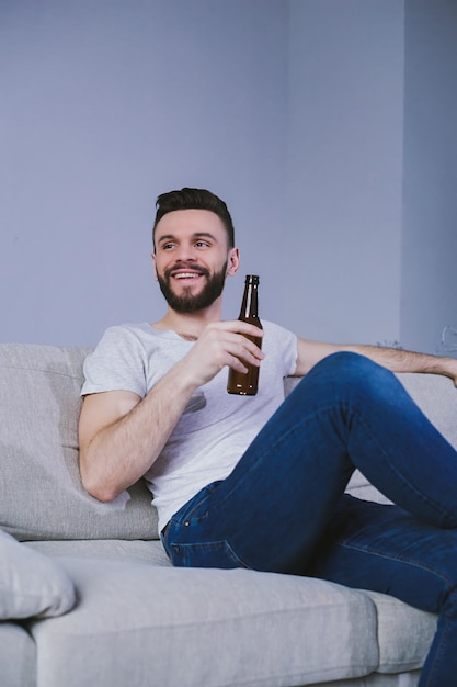 Bel homme barbu souriant regardant un match de football à la télévision assis à la maison sur le canapé et buvant de la bière