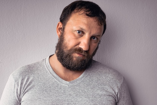 Un bel homme barbu en t-shirt gris regarde la caméra en se tenant debout contre un mur gris.