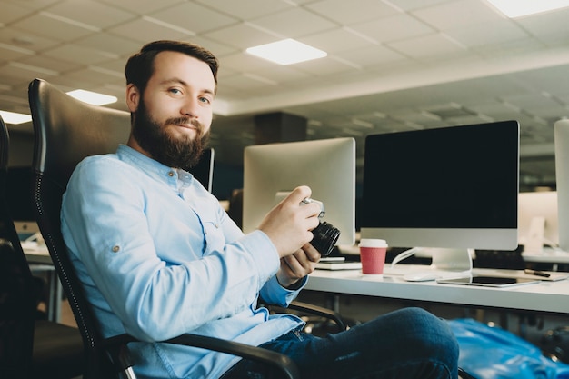 Bel homme barbu tenant la caméra et souriant alors qu'il était assis au bureau au bureau