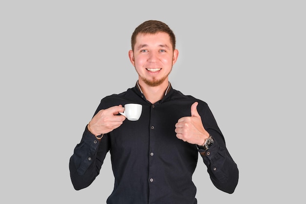 Un bel homme barbu tient une petite tasse de café dans ses mains et montre un geste du pouce levé en se tenant debout sur un fond gris