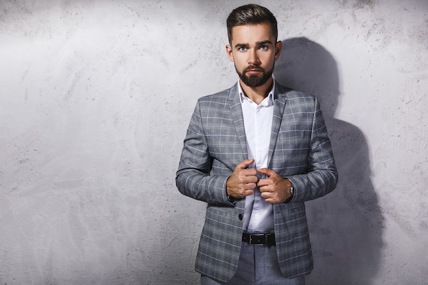 Bel homme barbu vêtu d'un costume à carreaux gris pose contre le mur de béton