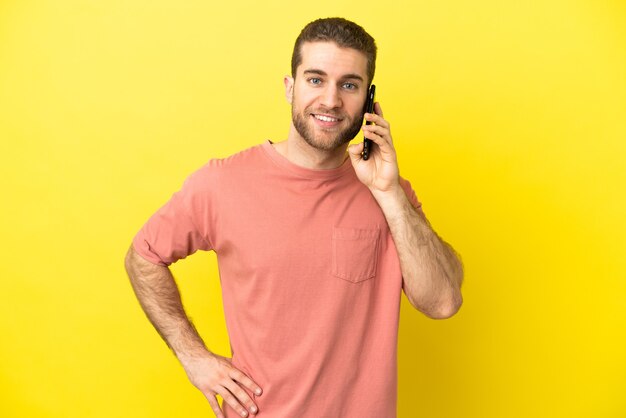 Bel homme blond à l'aide de téléphone mobile sur fond isolé posant avec les bras à la hanche et souriant