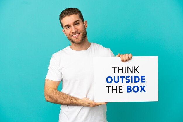 Bel homme blond sur fond bleu isolé tenant une pancarte avec texte Think Outside The Box avec une expression heureuse