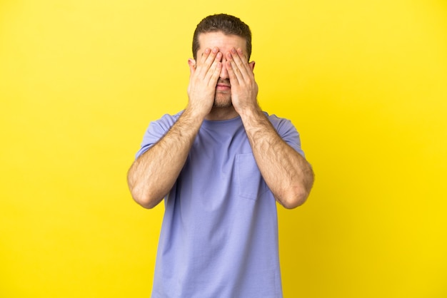 Bel homme blond sur fond jaune isolé couvrant les yeux par les mains