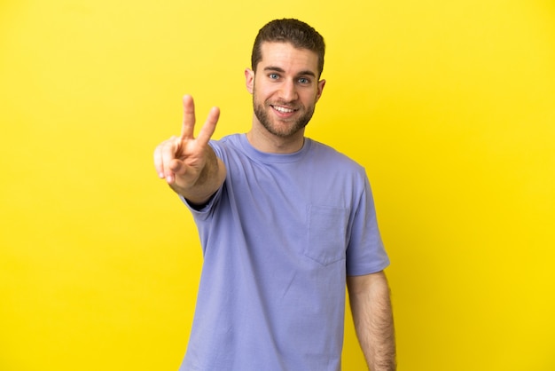 Bel homme blond sur fond jaune isolé souriant et montrant le signe de la victoire