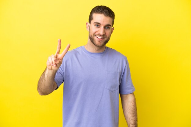 Bel homme blond sur fond jaune isolé souriant et montrant le signe de la victoire